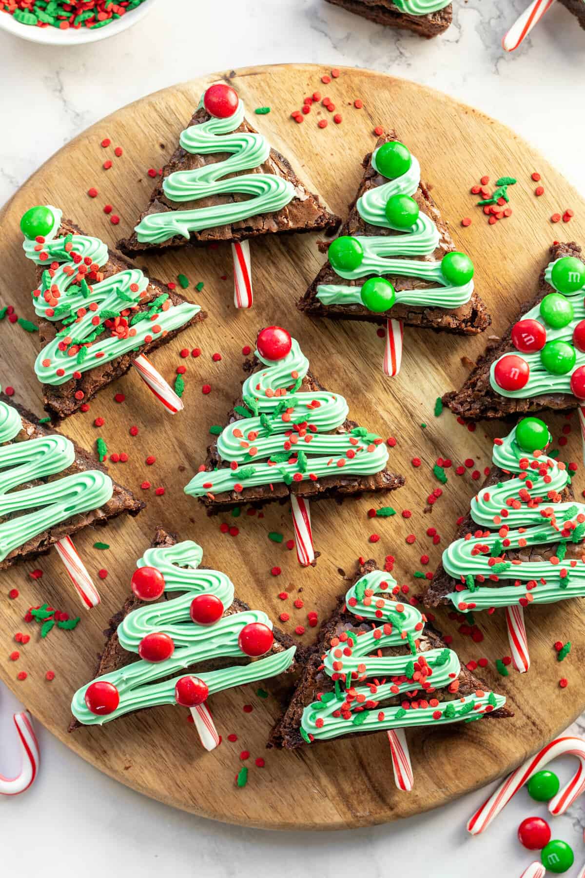 top down image of christmas tree brownies with garland green frosting and red and green sprinkles with M&Ms and candy cane sticks for decoration, served on a wooden round board