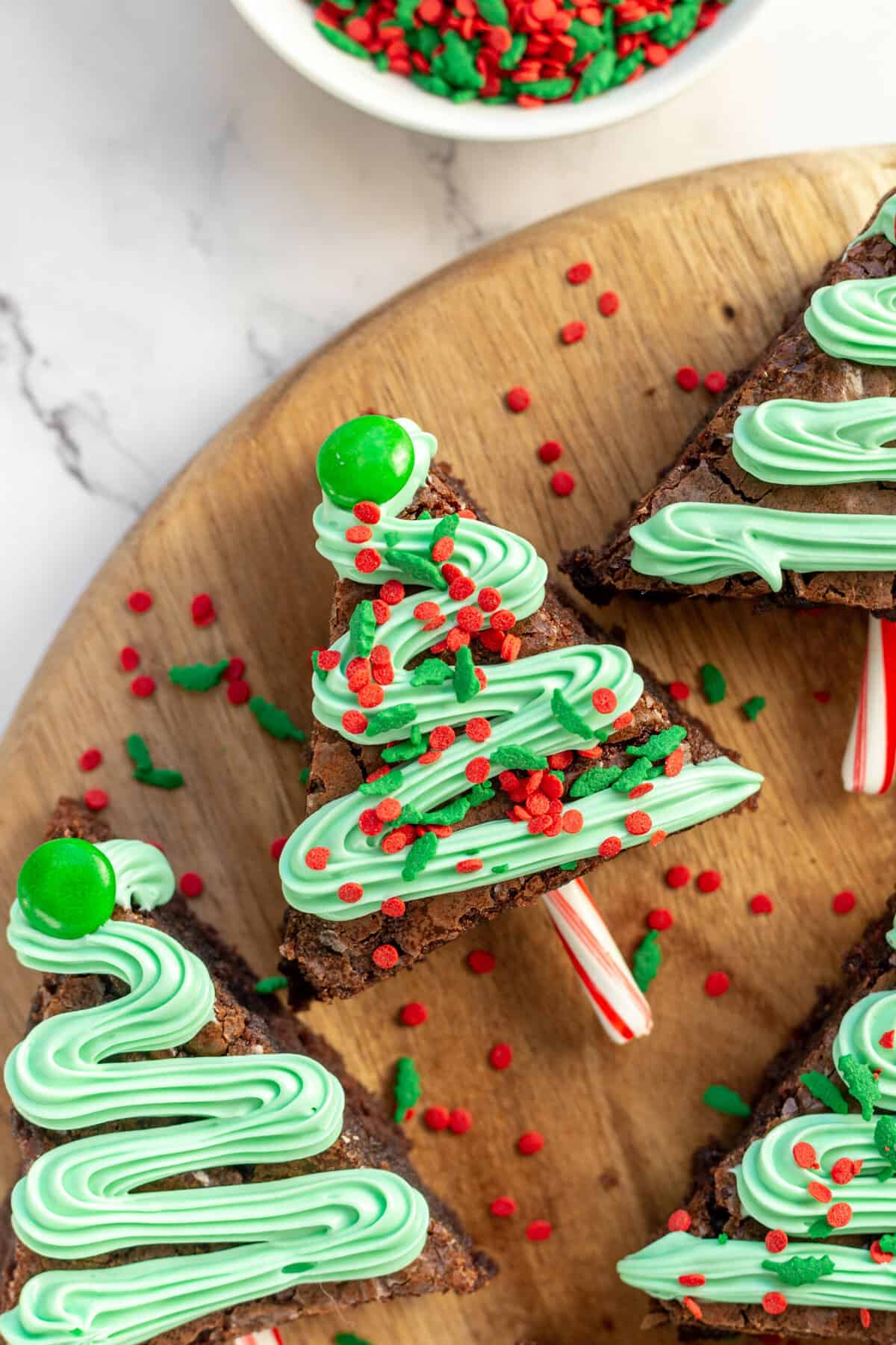 A close up image of a Christmas tree brownie with a frosting garland and red and green sprinkles with a green M&M for a star and a candy cane stick for a trunk, served on a round wooden board.