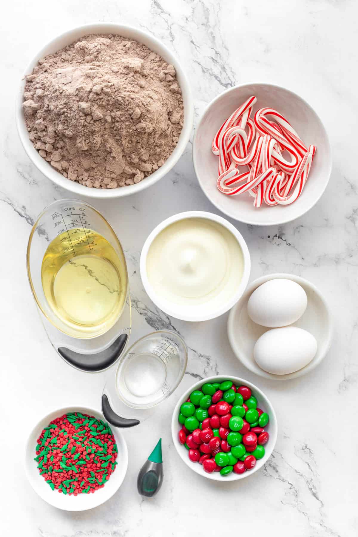 The ingredients to make Christmas tree brownies are shown portioned out on a white background: brownie mix, oil, sour cream, eggs, candy canes, and candy.