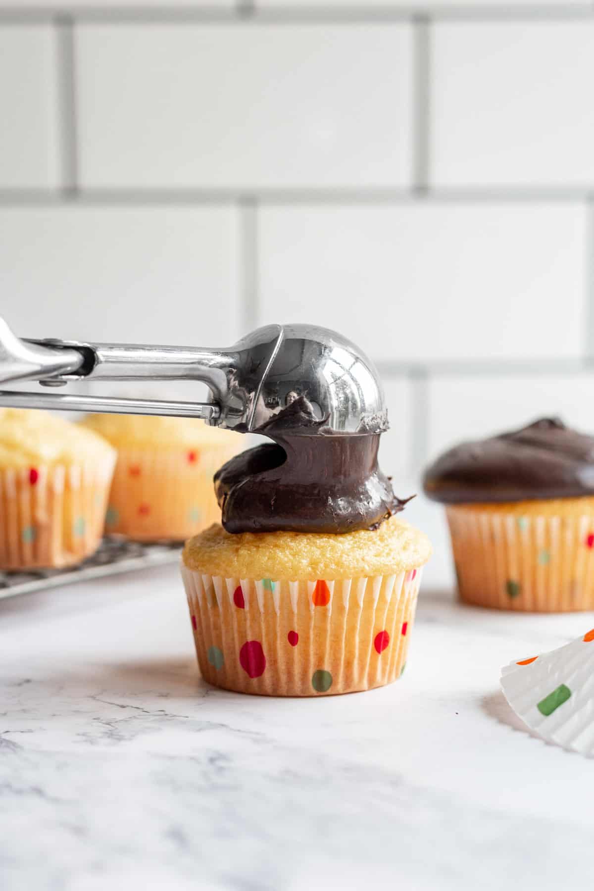 using an ice cream scoop to frost chocolate frosting on a vanilla cupcake