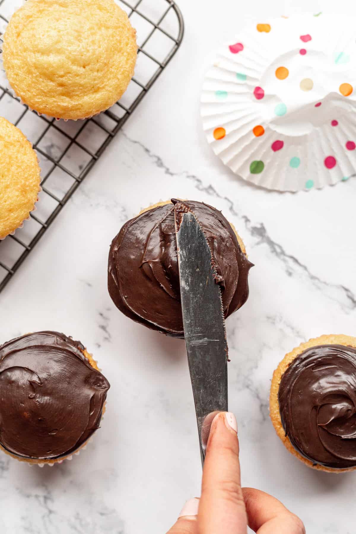 using a knife to spread chocolate frosting onto vanilla cupcakes