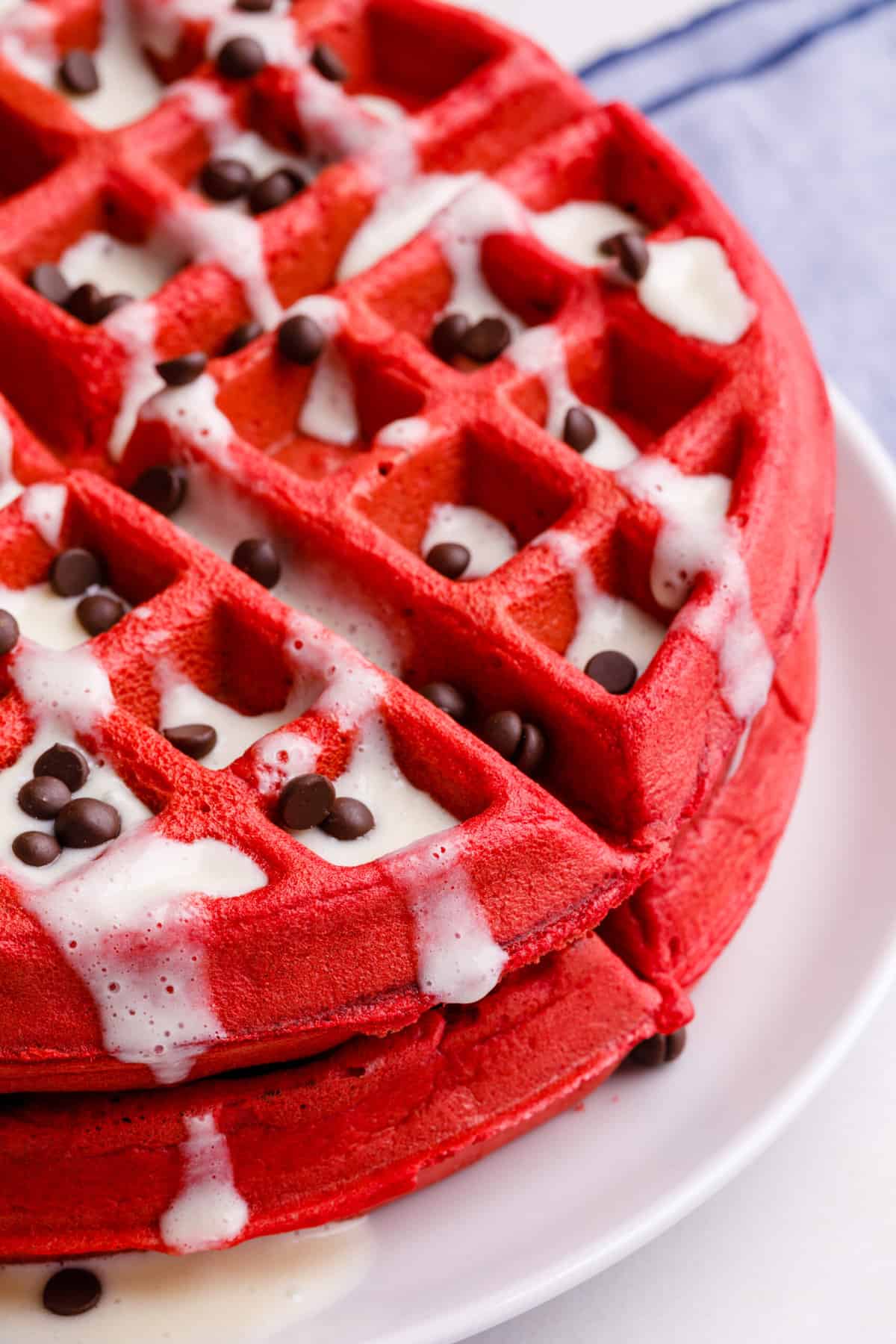 close up image of a stack of red velvet waffles with cream cheese glaze topped with mini chocolate chips served on a white round plate