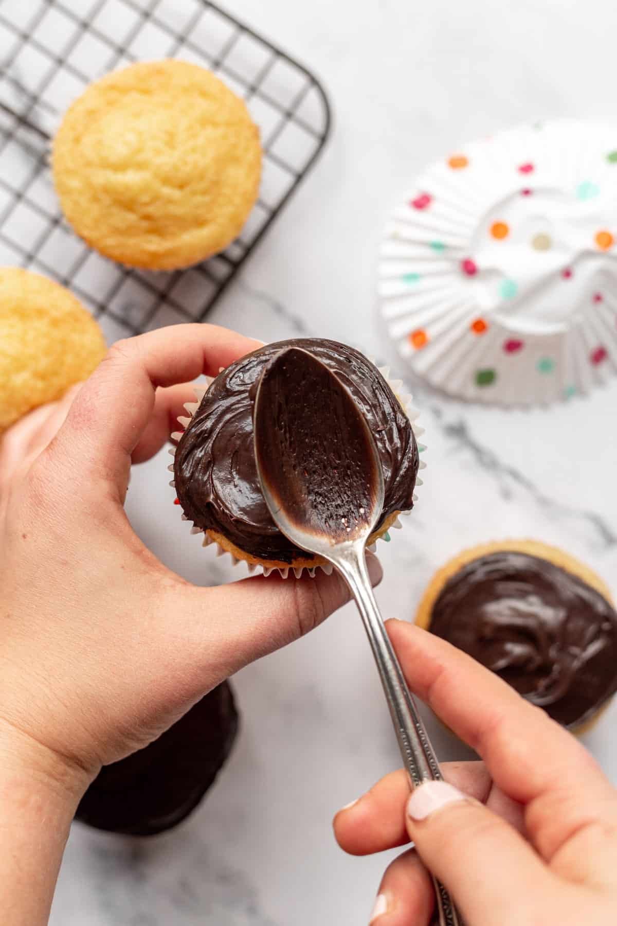 using a spoon to spread chocolate frosting on a vanilla cupcake