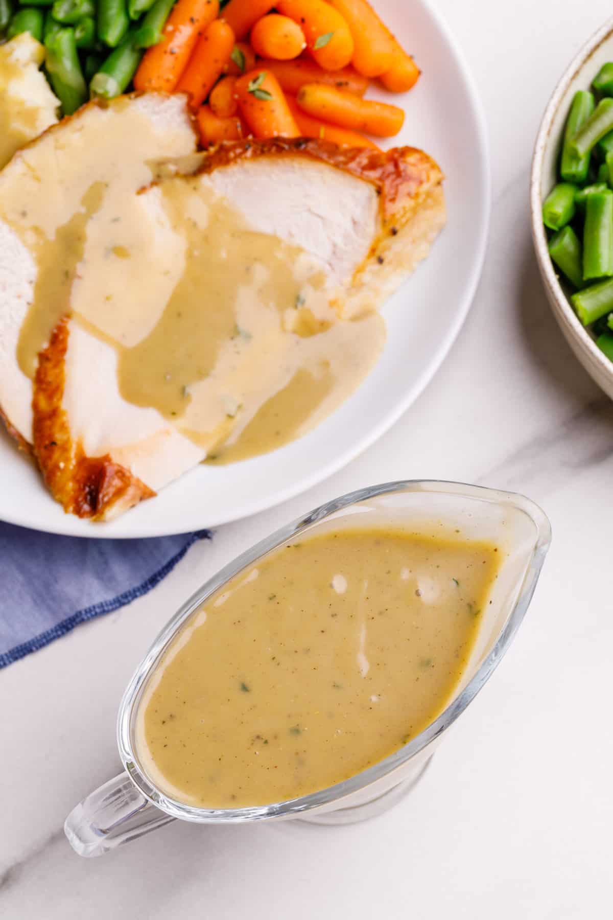 A top down image of turkey gravy in a clear gravy boat and a plate of Thanksgiving dishes.