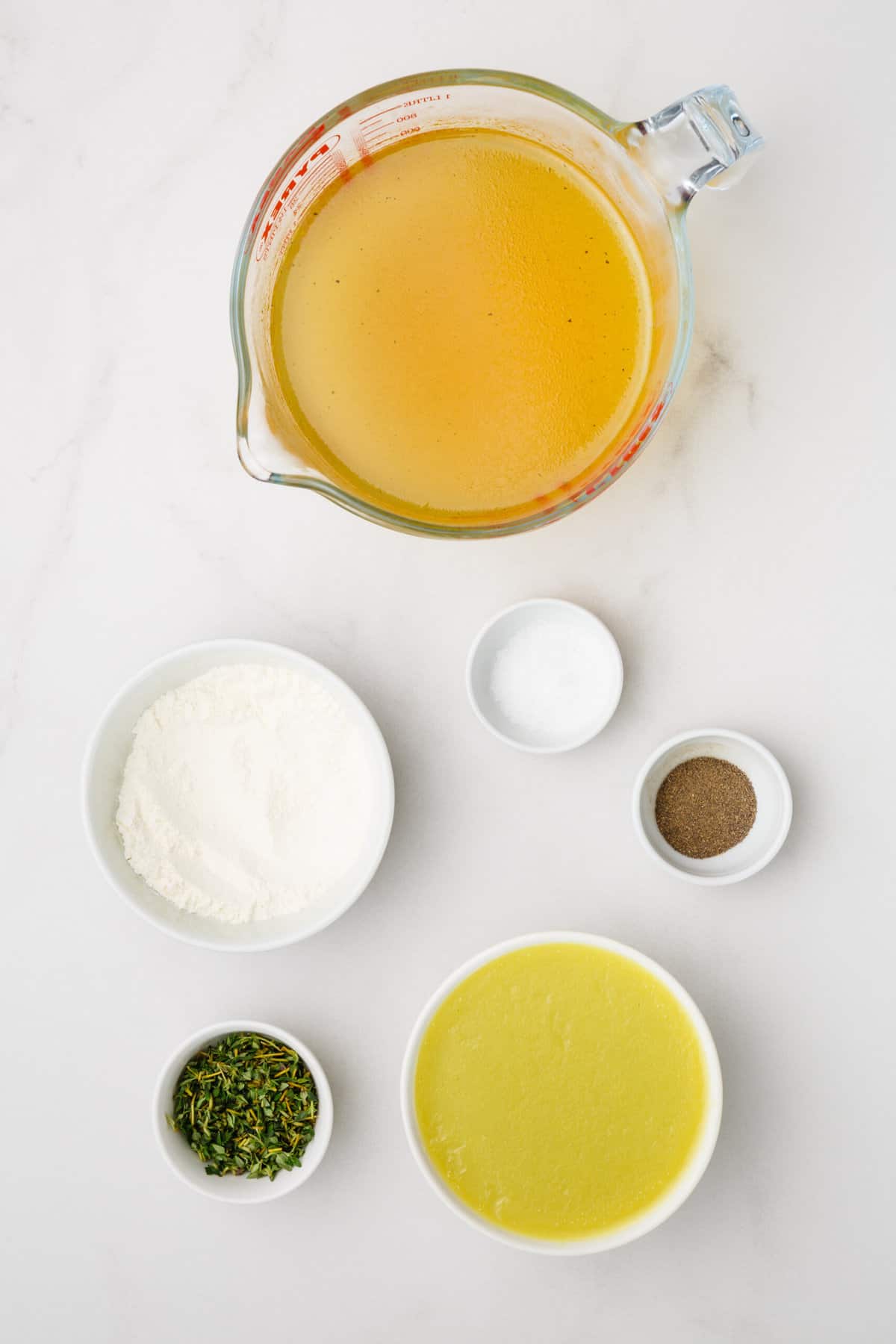The ingredients to make turkey gravy are shown portioned out on a white background: turkey broth, butter, flour, thyme, and salt and pepper.