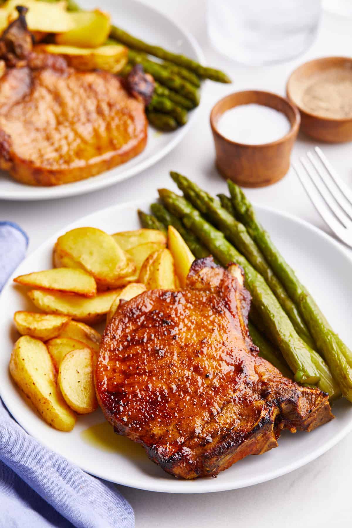 plate of air fryer bone in pork chops served with a side of fried potato wedges and asparagus sitting on a white round plate.