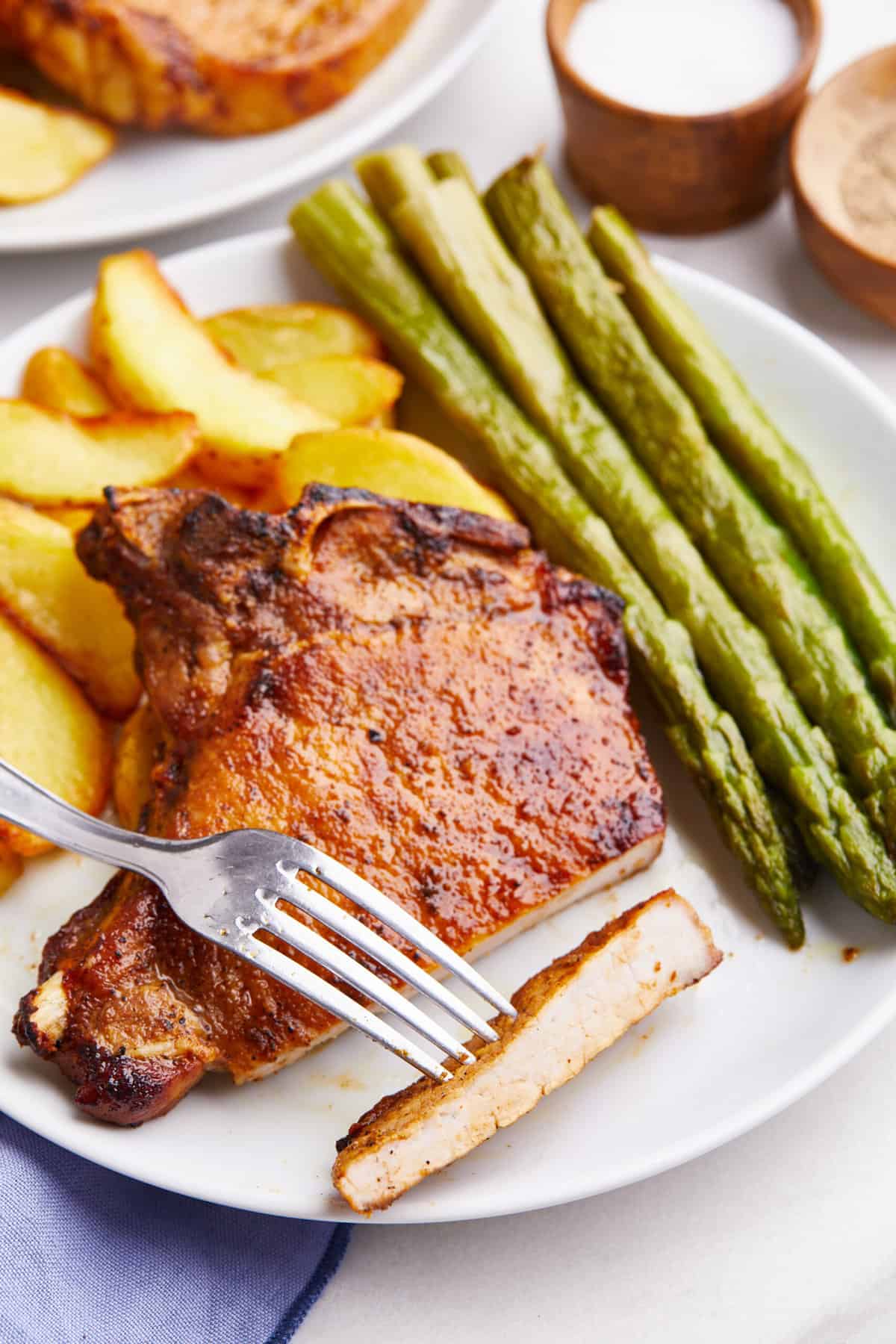 close up image of a plate of air fryer bone in pork chops with a side of fried potato wedges and asparagus cut in half to show the cross section.