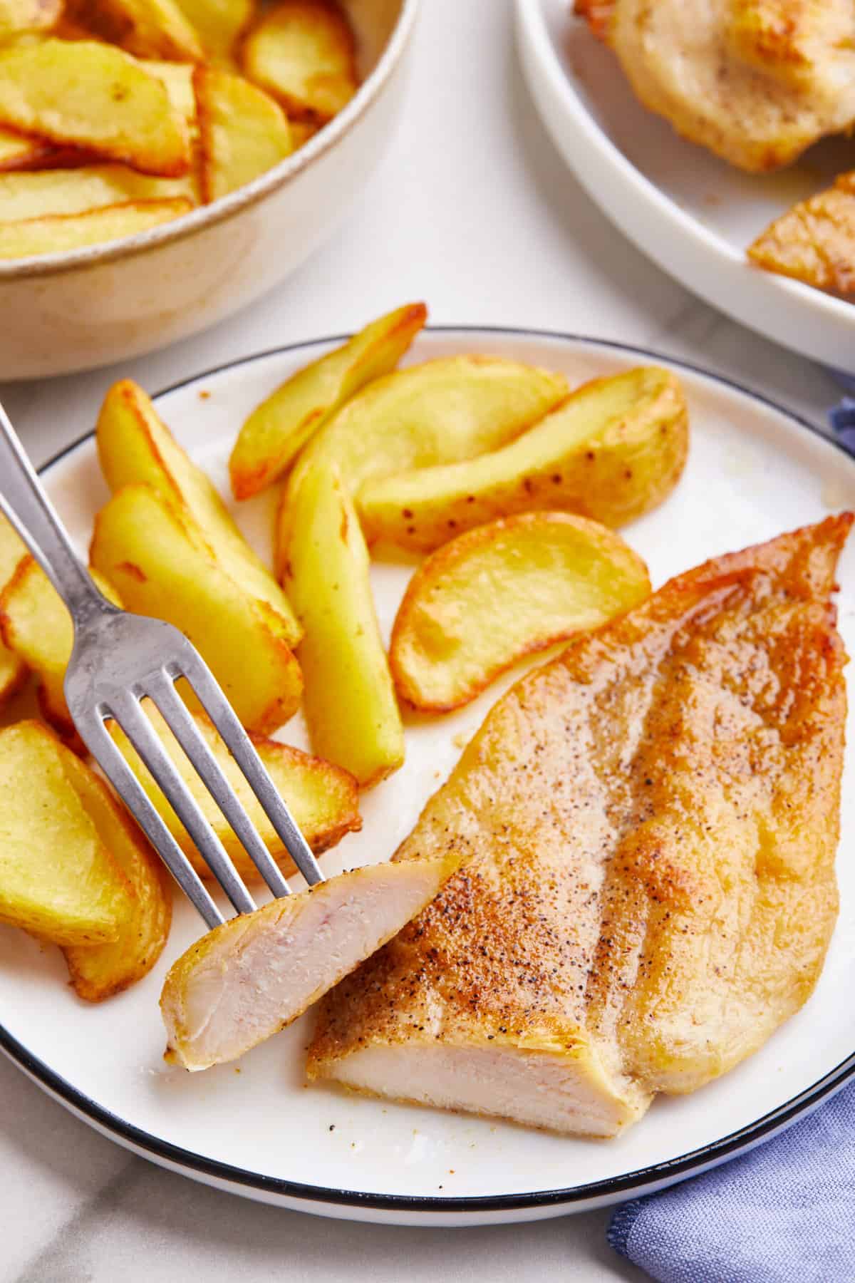 broiled chicken breast cut with a knife to show the cross section served on a white round plate with a side of fried potato wedges