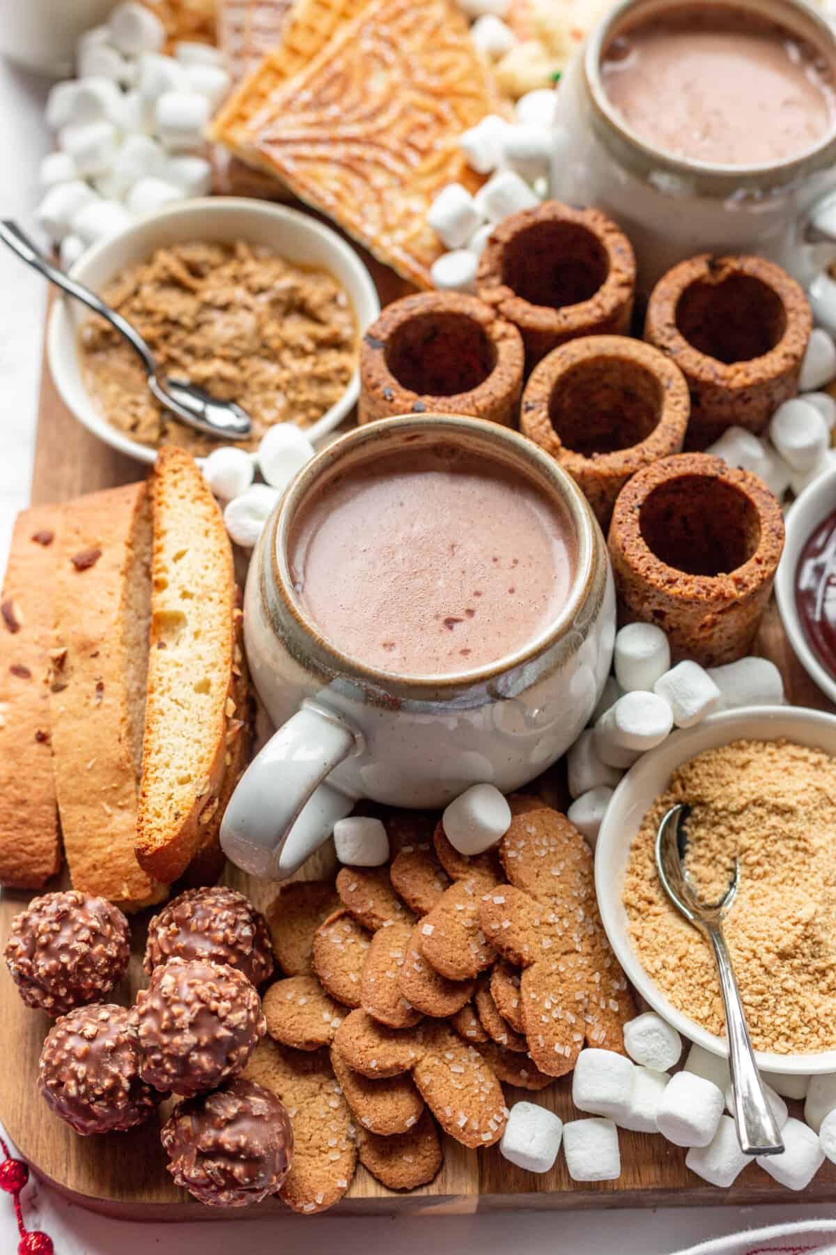 Mugs of hot chocolate surrounded by cookies and toppings. 
