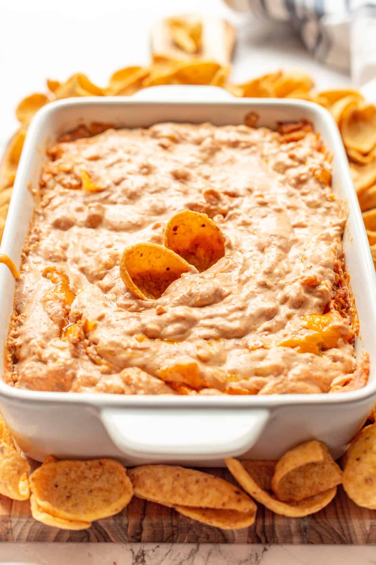 close up image of creamy and cheesy refried bean dip with two fritos sitting in a large white casserole dish surrounded by frito chips