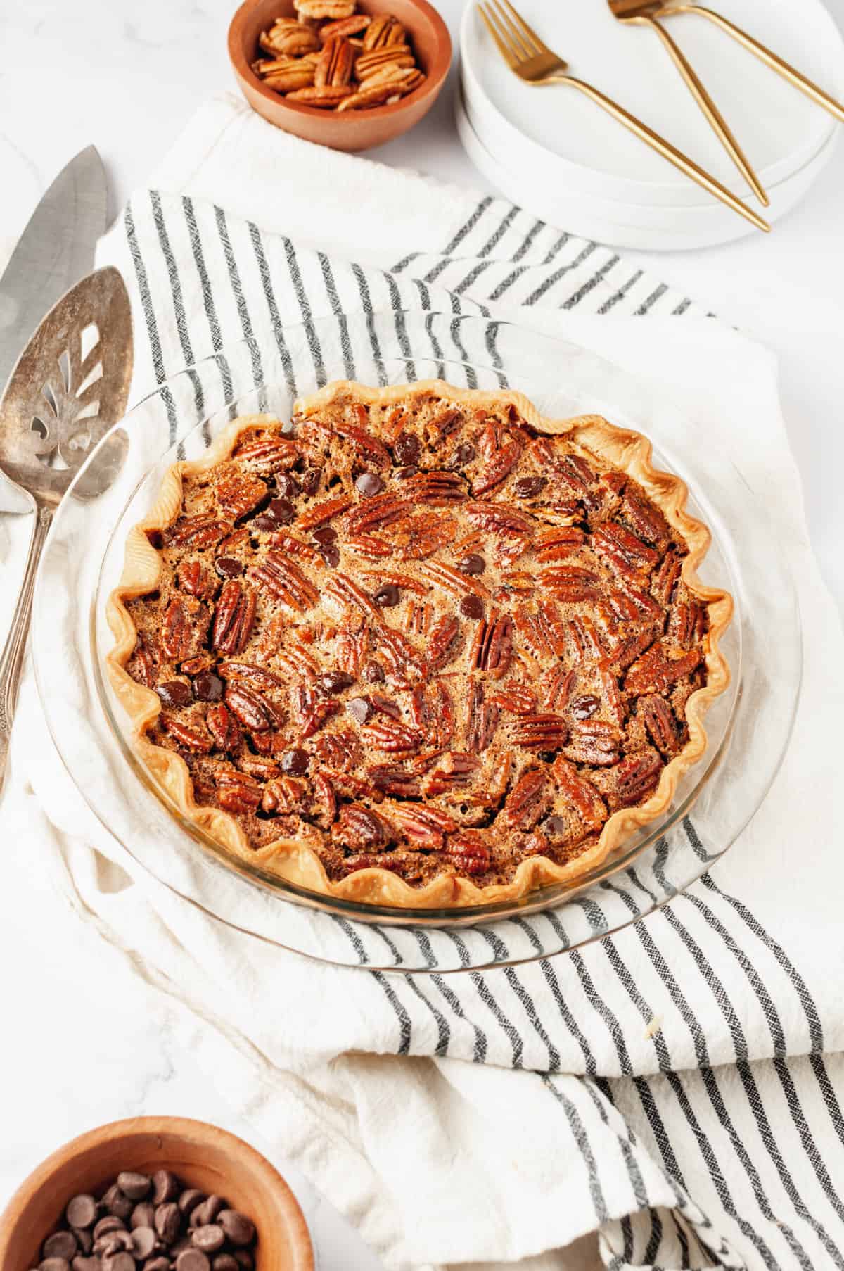 Top down image of a finished baked chocolate pecan pie served in a clear pie dish, striped kitchen cloth, silver server, small bowl of pecans, white serving plates and forks in the background.