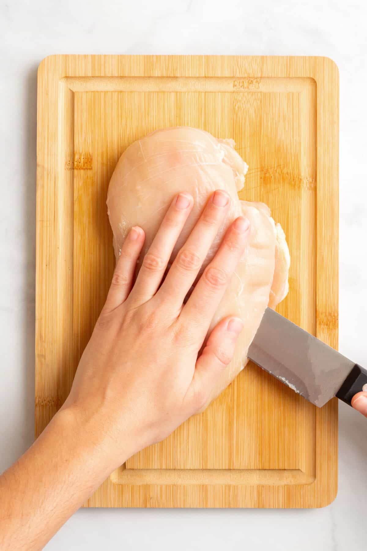 A hand holding raw chicken on a cutting board while a second hand slices it. 