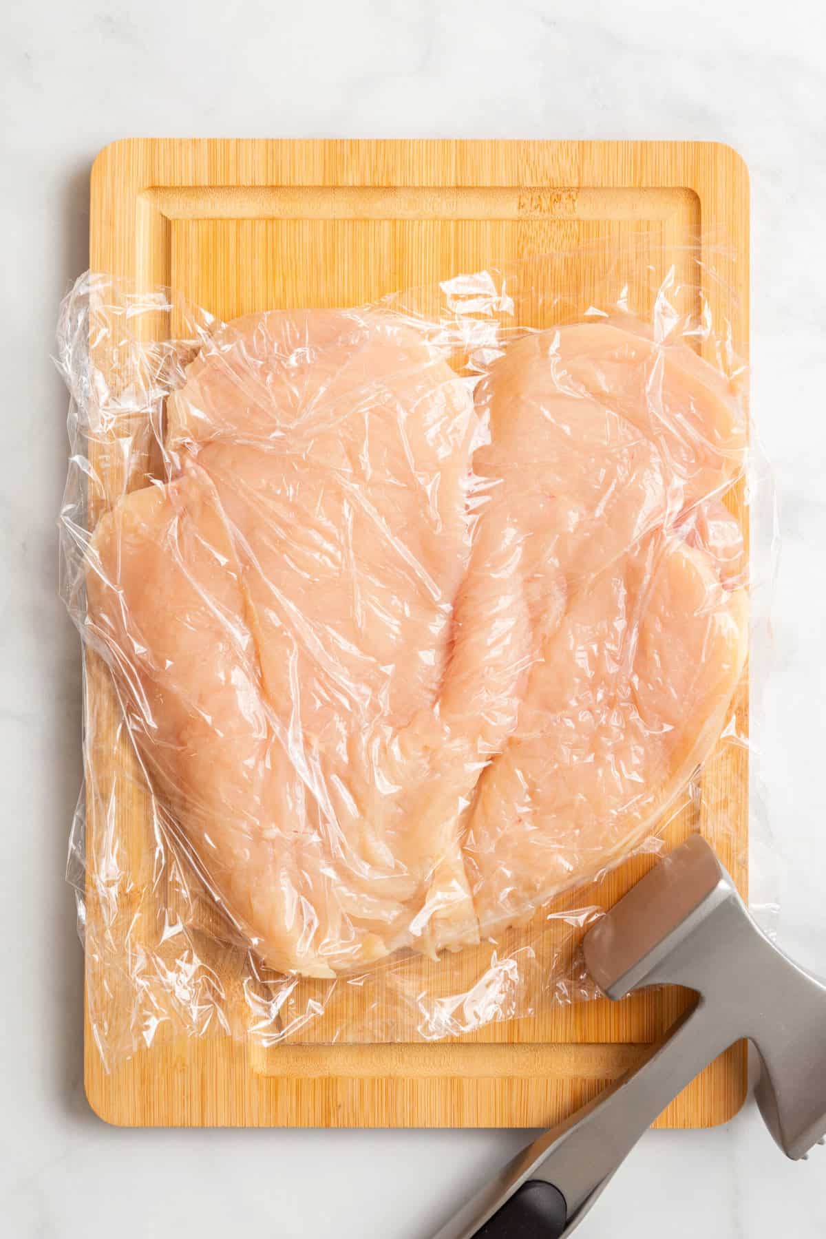 pounded butterflied chicken breast wrapped in cling wrap sitting on a wood cutting board. 