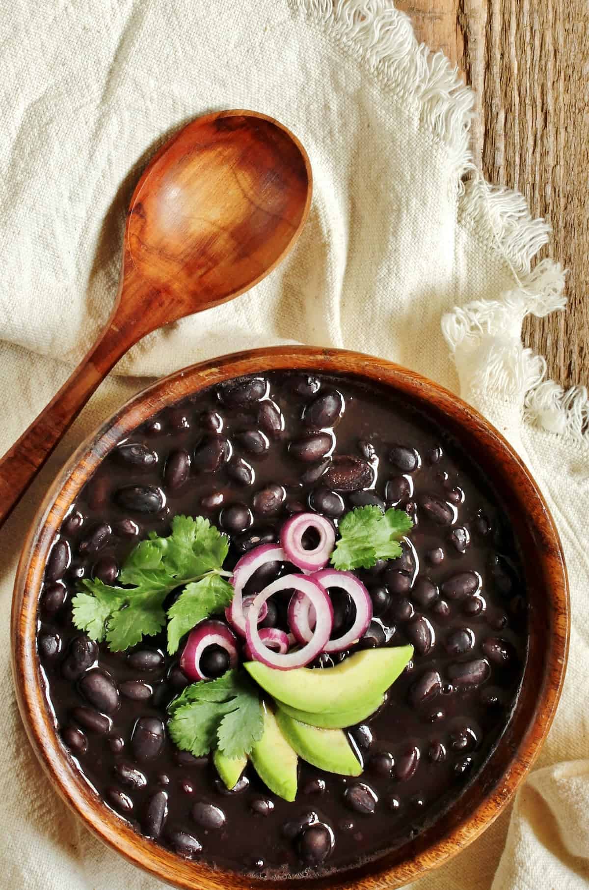 wooden bowl of instant pot black beans topped with sliced avocados, red onion and cilantro