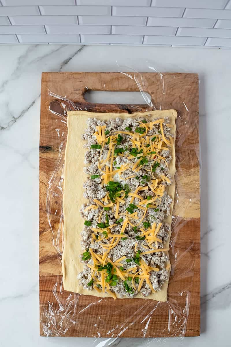 Cheese and chives are placed on top of sausage filling and dough on a cutting board.