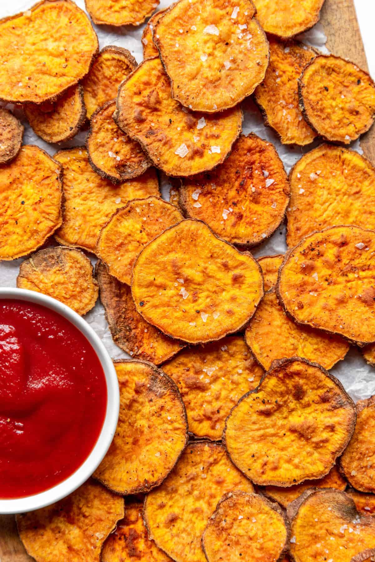 top down close up image of air fryer sweet potato chips sitting on parchment paper with a side of ketchup in a white round bowl.