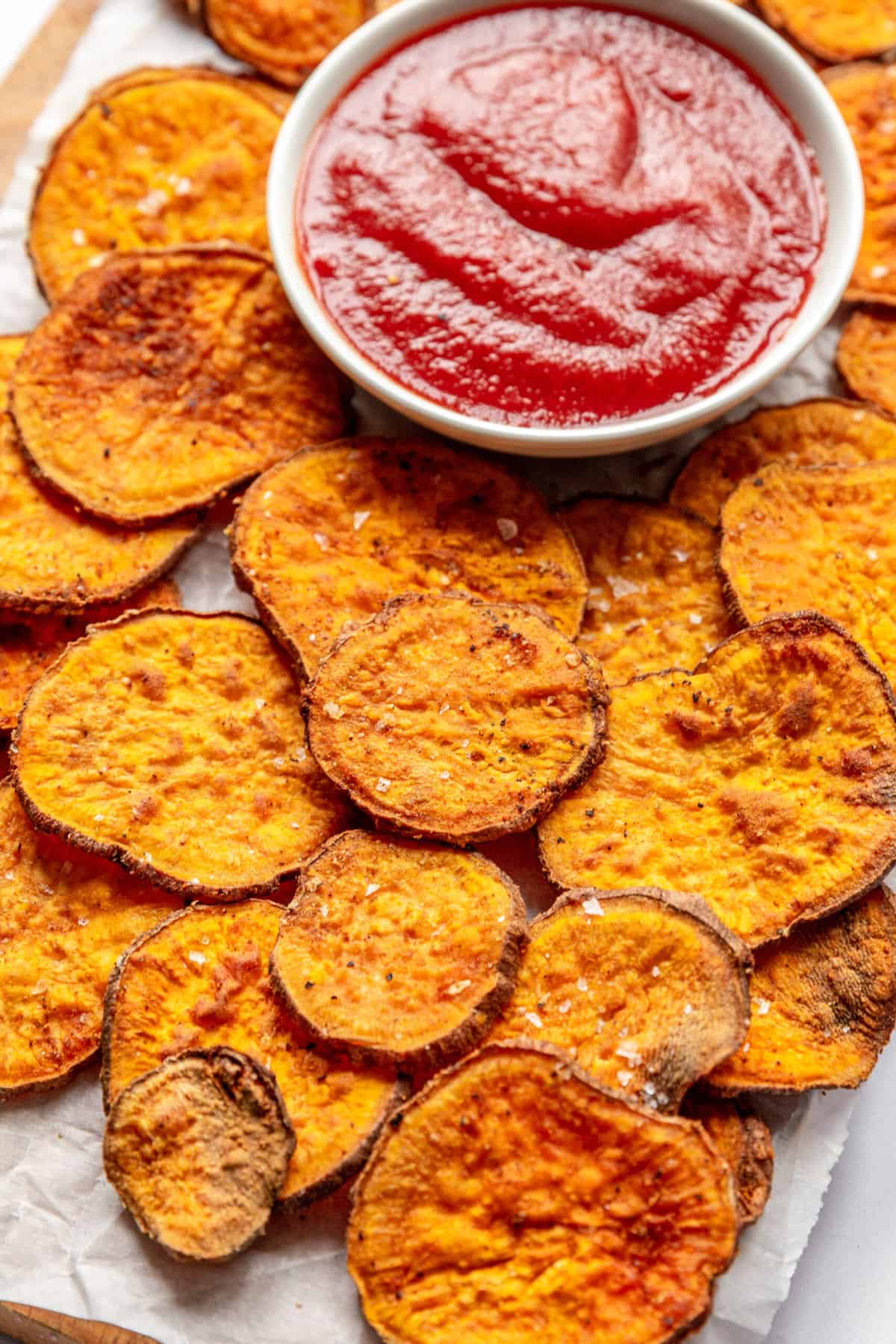 close up image of air fryer sweet potato chips sitting on parchment paper with a side of ketchup in a white round bowl.