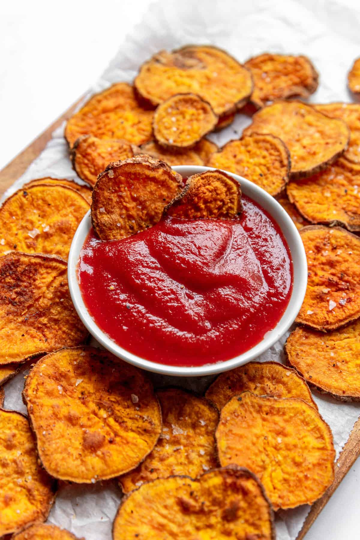 air fryer sweet potato chips sitting on parchment paper with a bowl of ketchup in the center.