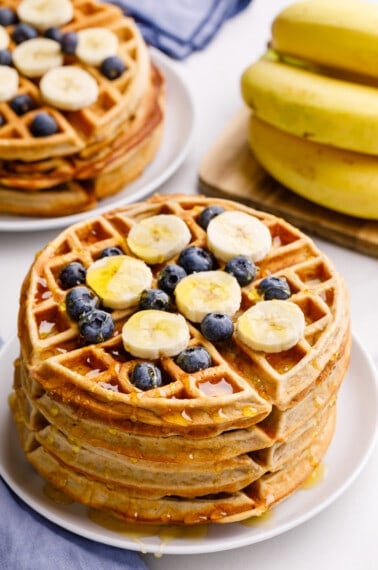 A stack of banana waffles topped with banana coins, blueberries, and syrup.