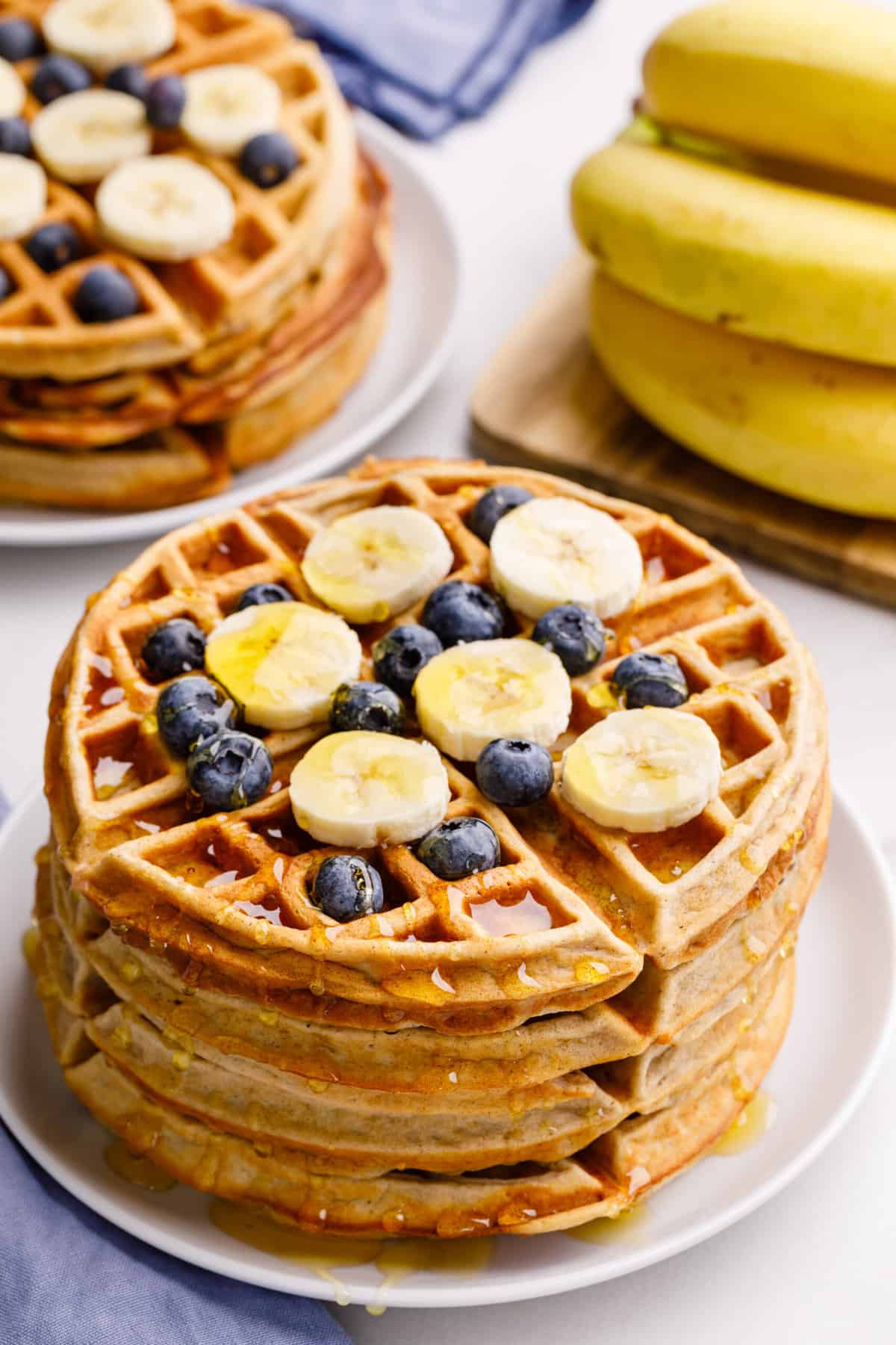 stack of four banana waffles topped with fresh sliced bananas and blueberries with syrup served on a white round plate.