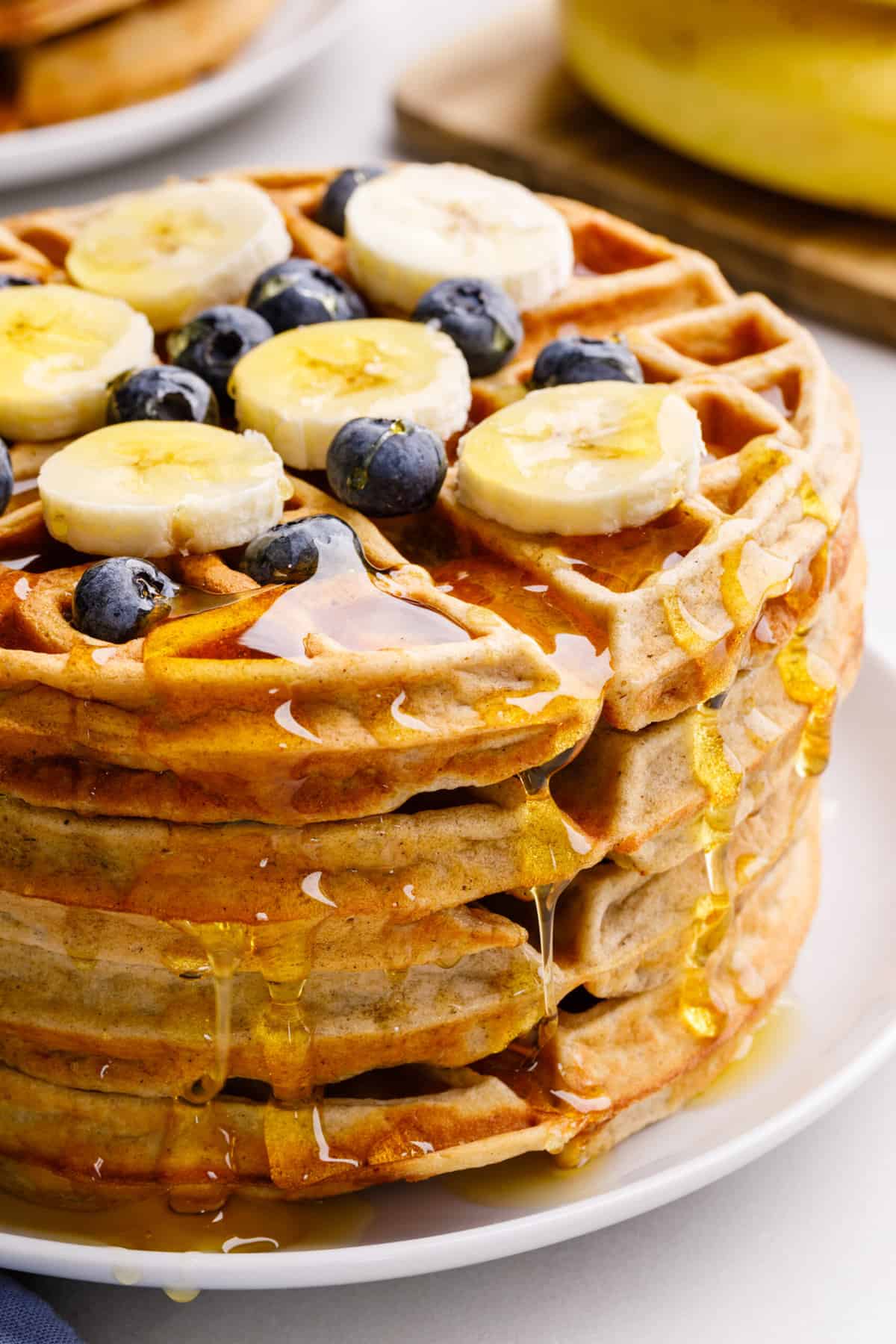 close up image of a stack of four banana waffles topped with syrup, sliced bananas and fresh blueberries served on a white round plate