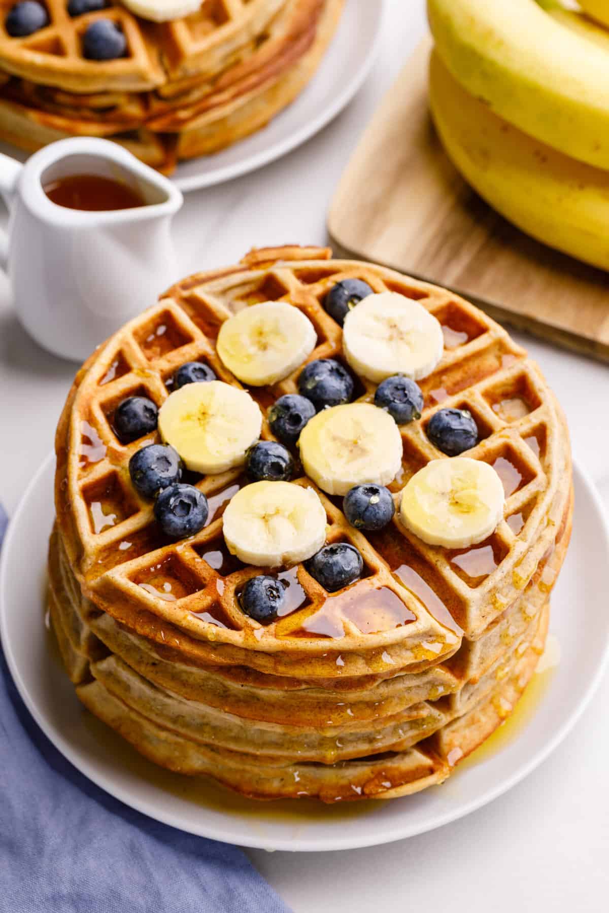 stack of four banana waffles topped with fresh blueberries, sliced bananas and syrup served on a white round plate.