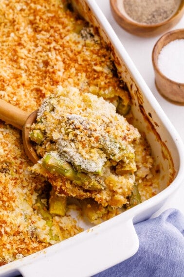 A spoon lifting a bite of cheesy asparagus casserole from a baking dish.