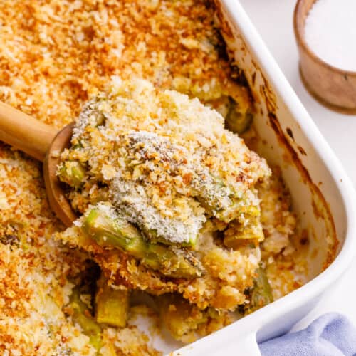 A spoon lifting a bite of cheesy asparagus casserole from a baking dish.