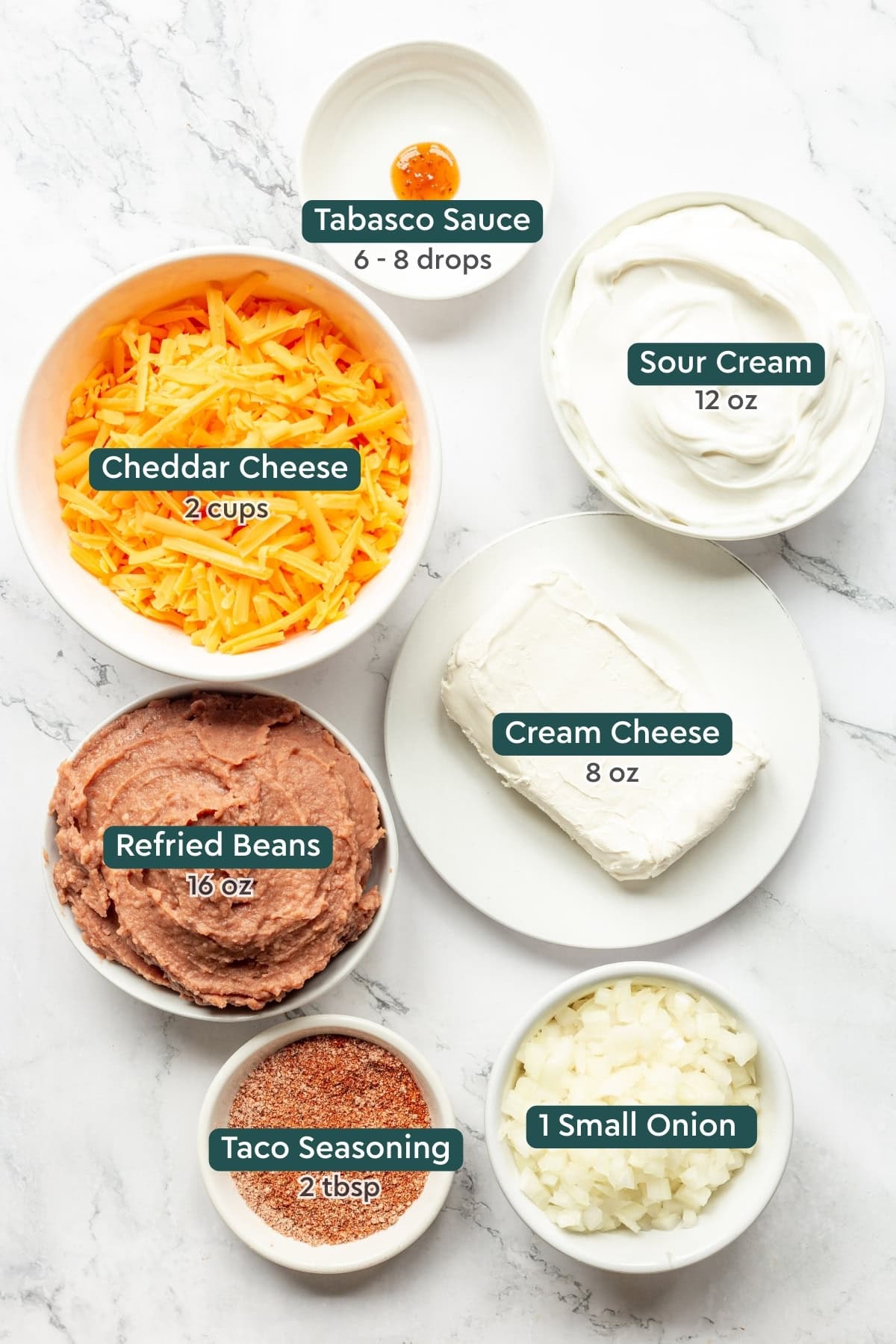 Ingredients for cheesy refried bean dip, including tabasco, sour cream, cheddar, cream cheese, refried beans, taco seasoning, onions.