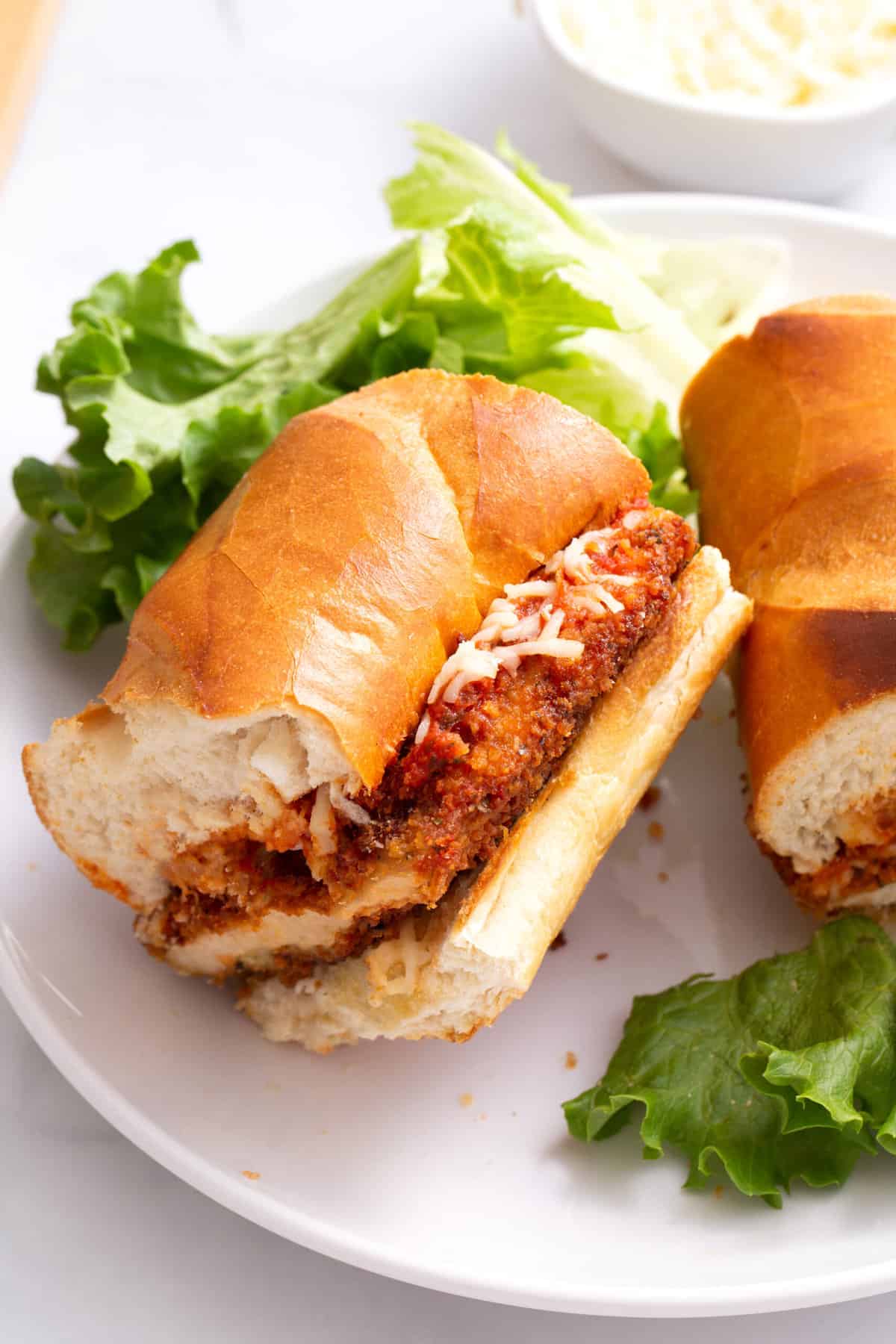 close up image of a chicken parmesan sandwich served on a white round plate with a bed of lettuce.