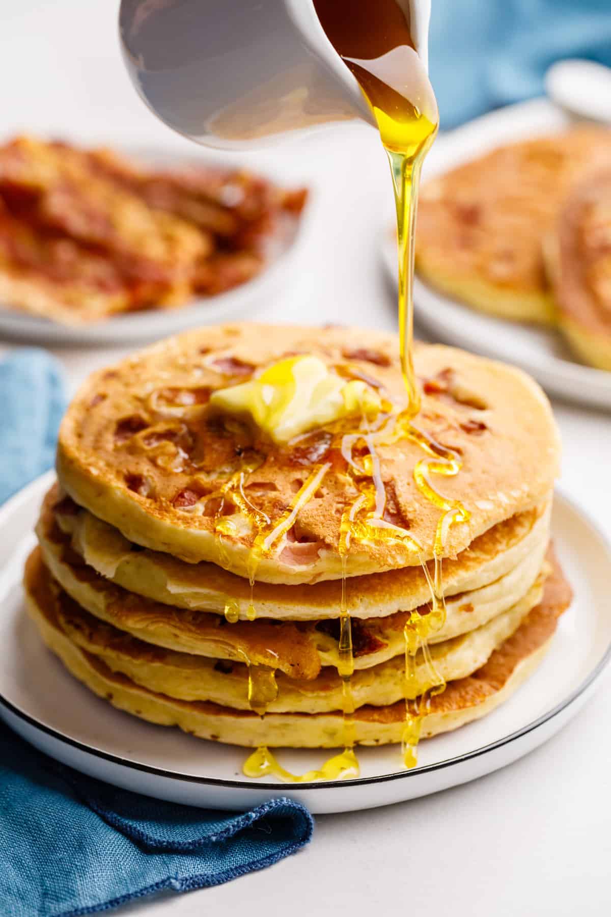 maple syrup being pour on top of a stack of five bacon pancakes served on a white round plate