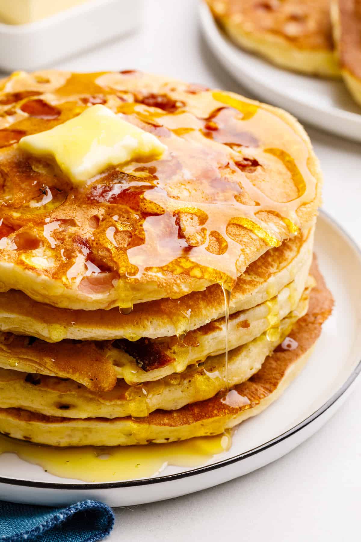 close up image of a stack of five bacon pancakes topped and ripping with maple syrup down the sides with a pad of butter on top