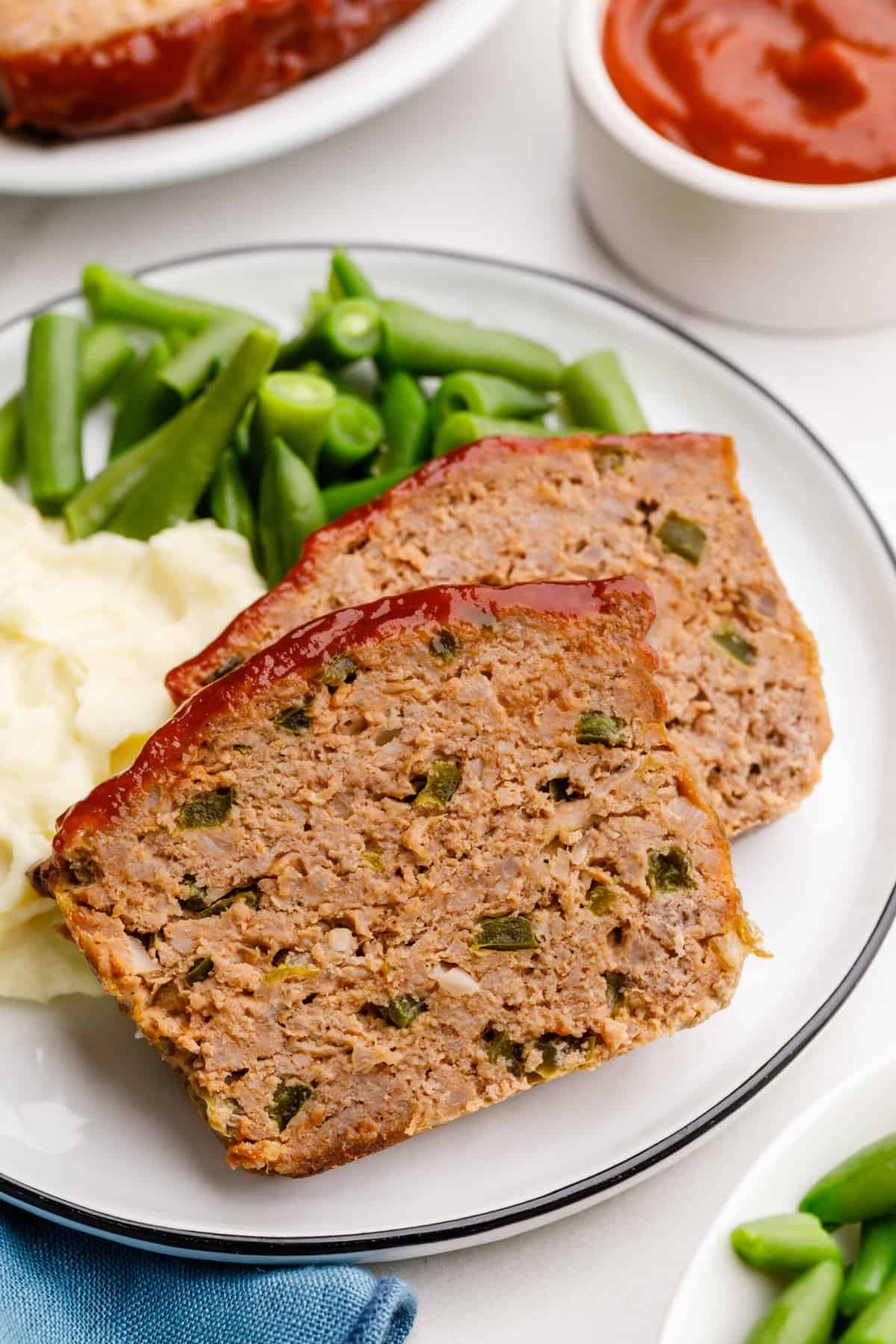 close up image of two servings of southern meatloaf served on a white round plate showing the cross section, served with a side of mashed potatoes and green beans