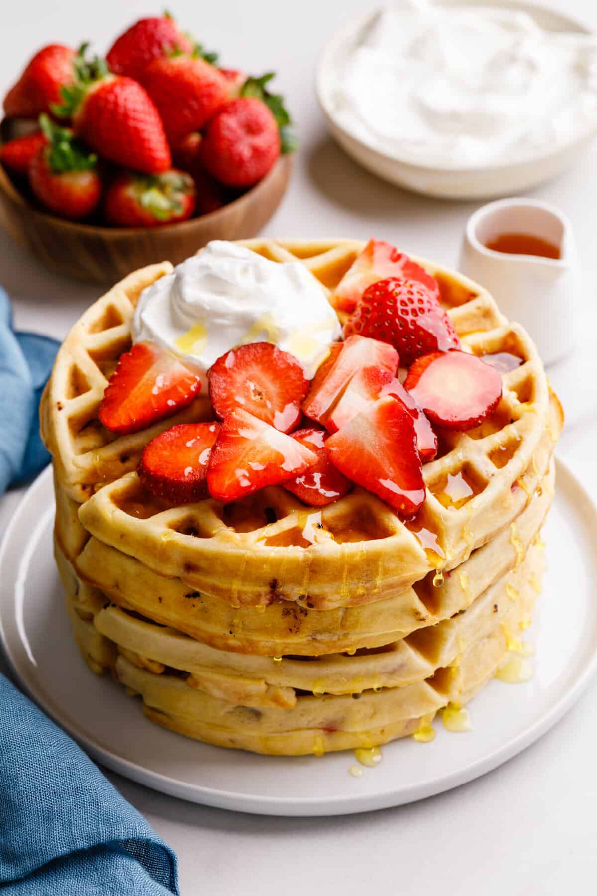 stack of four strawberry waffles topped with syrup, whipped cream and fresh strawberries, served on a white round plate