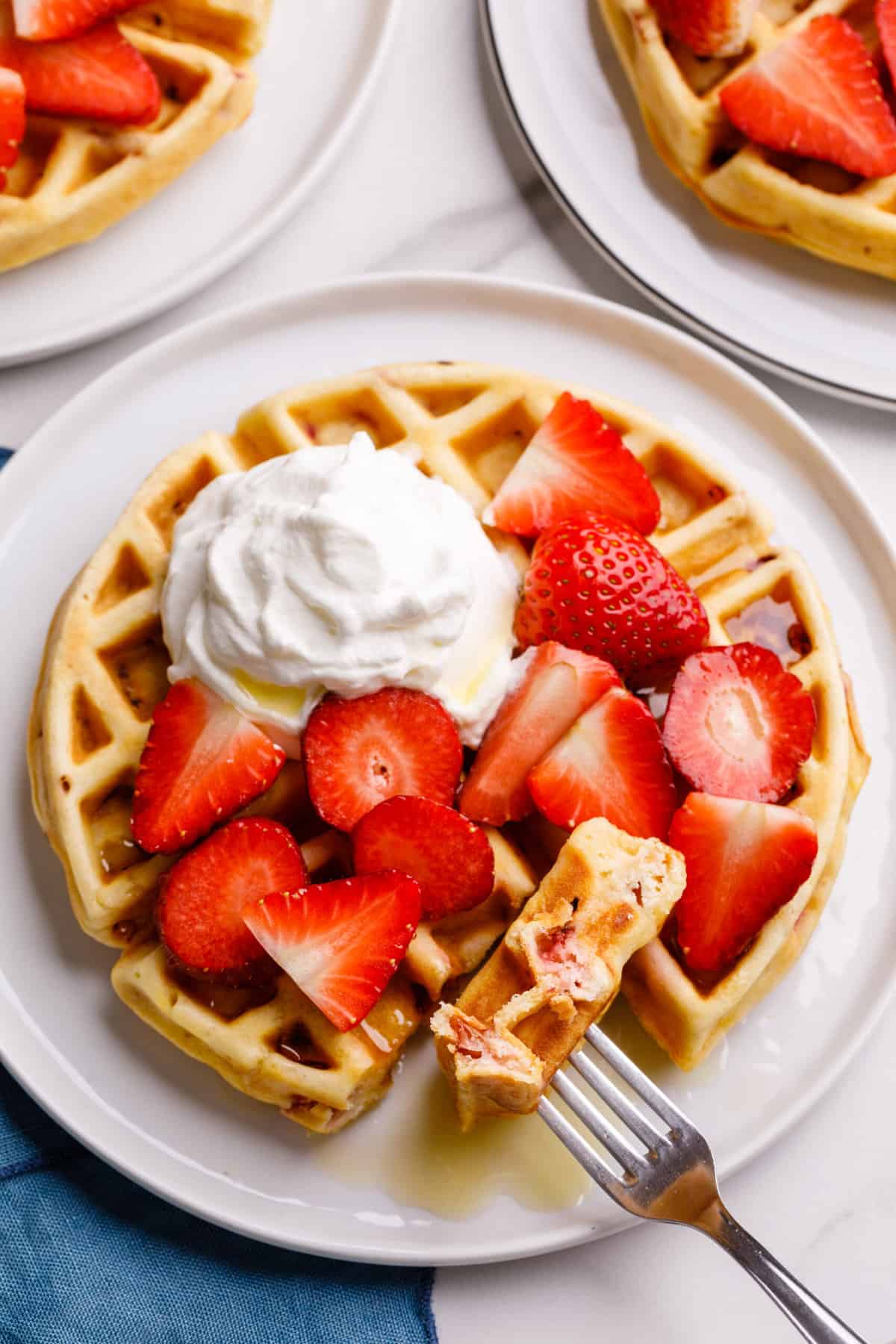 serving of strawberry waffles topped with whipped cream and fresh cut strawberries and syrup sitting on a white round plate