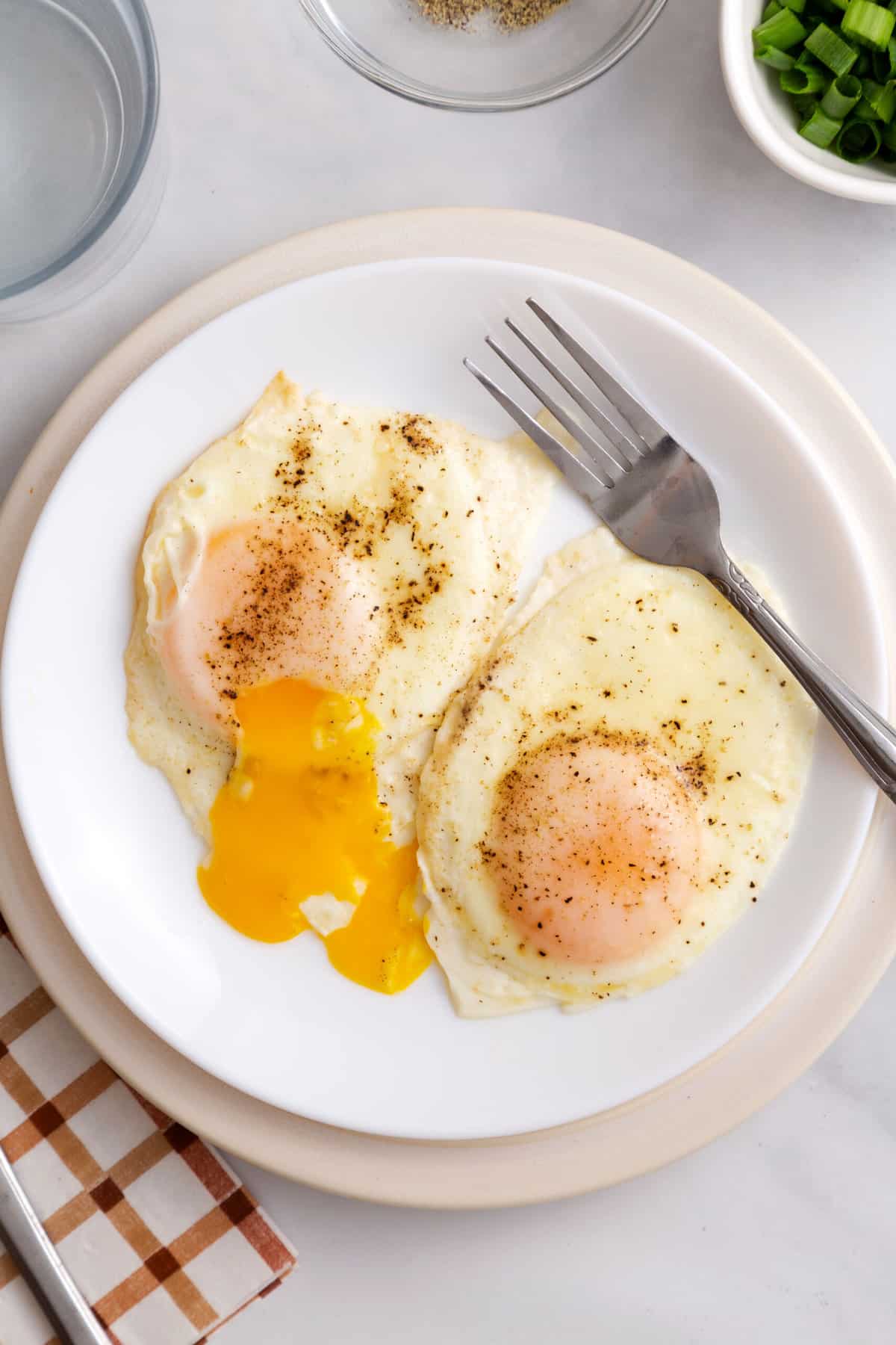 top down image of two basted eggs  with one cracked runny yolk served on a white round plate with a silver fork