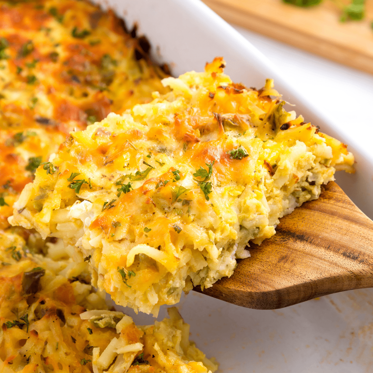 A slice of cheesy breakfast potato casserole being lifted from a casserole dish.