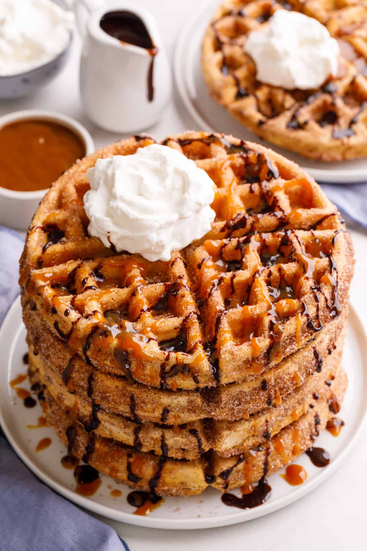 stack of four churro waffles on a plate topped with chocolate and caramel sauce and whipped cream