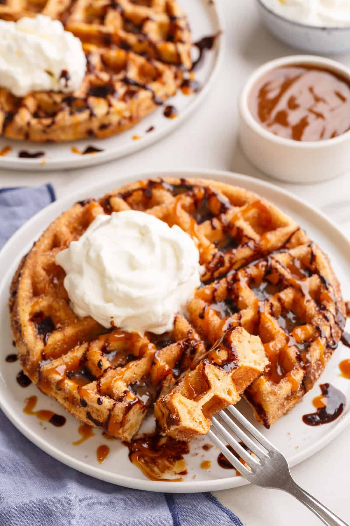 serving of churro waffle on a white round plate topped with chocolate sauce, dulche de leche and whipped cream