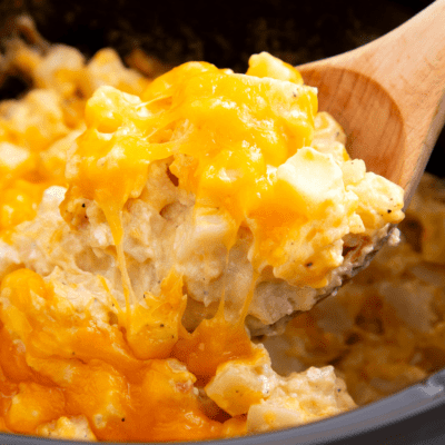 A wooden spoon lifting a scoop of cheesy potatoes from a crockpot.