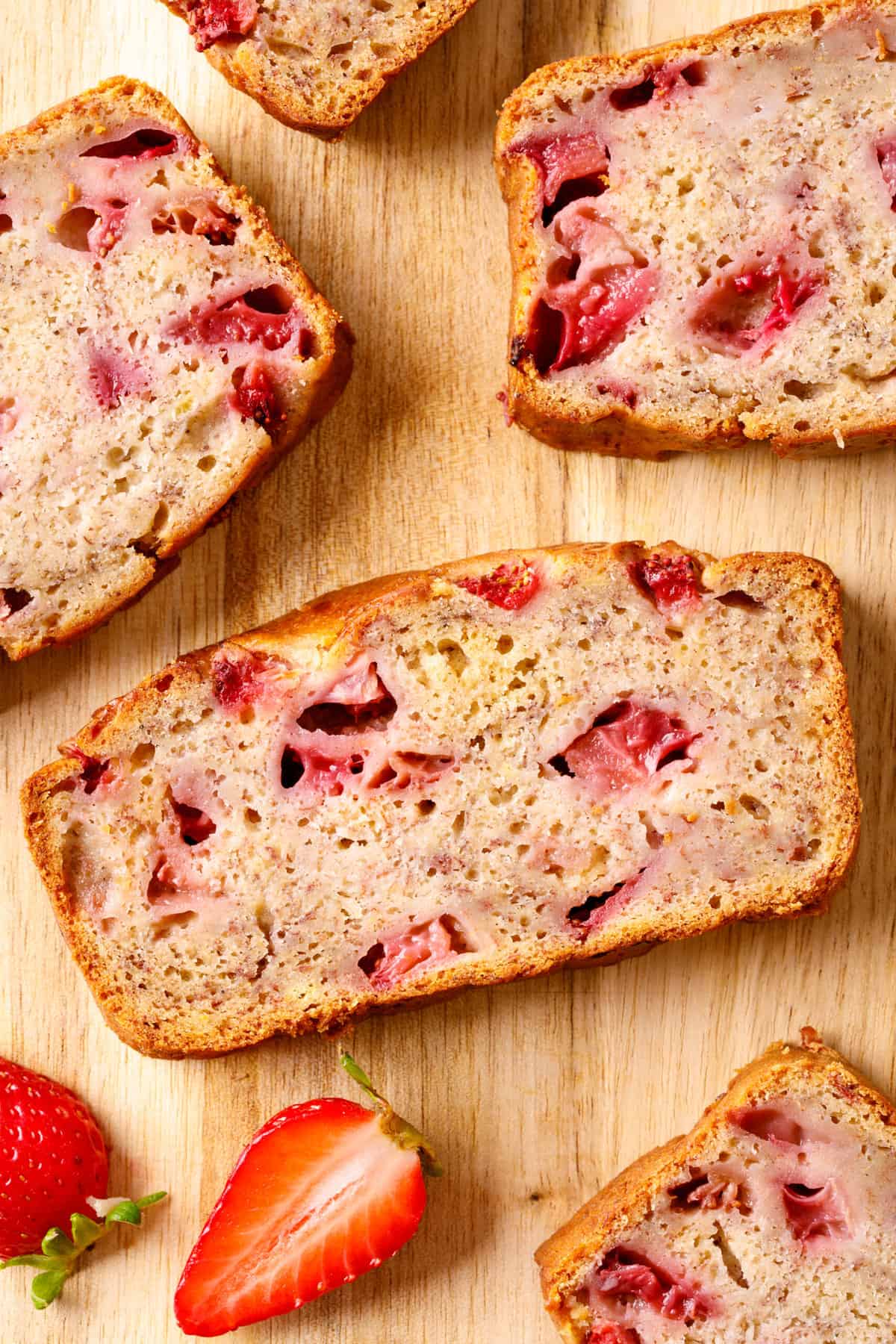 Top down image of slices of strawberry banana bread sitting on a wood cutting board with fresh cut strawberries surrounding.