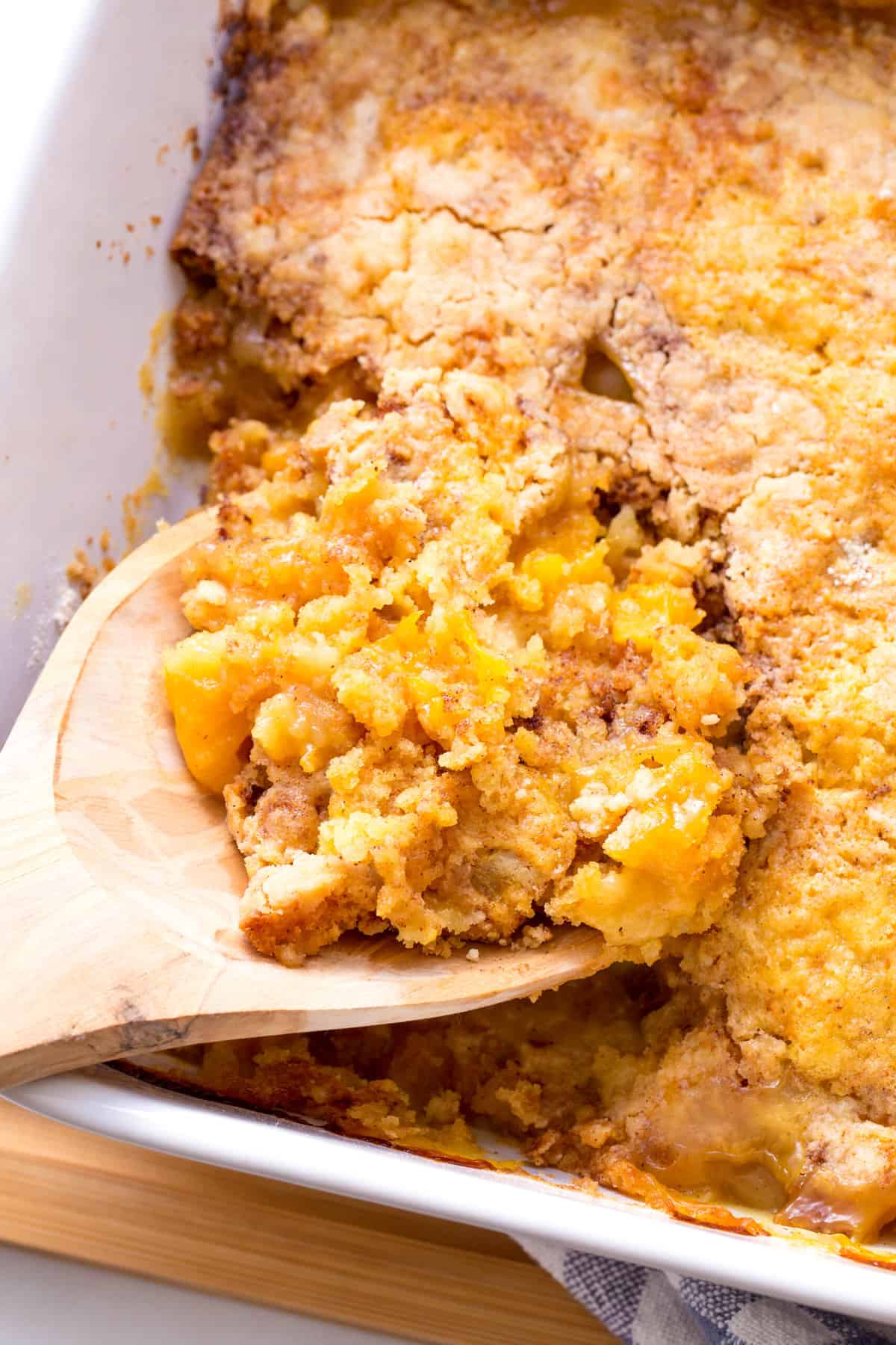 close up image of a wooden spoonful digging into a casserole dish of peach cobbler
