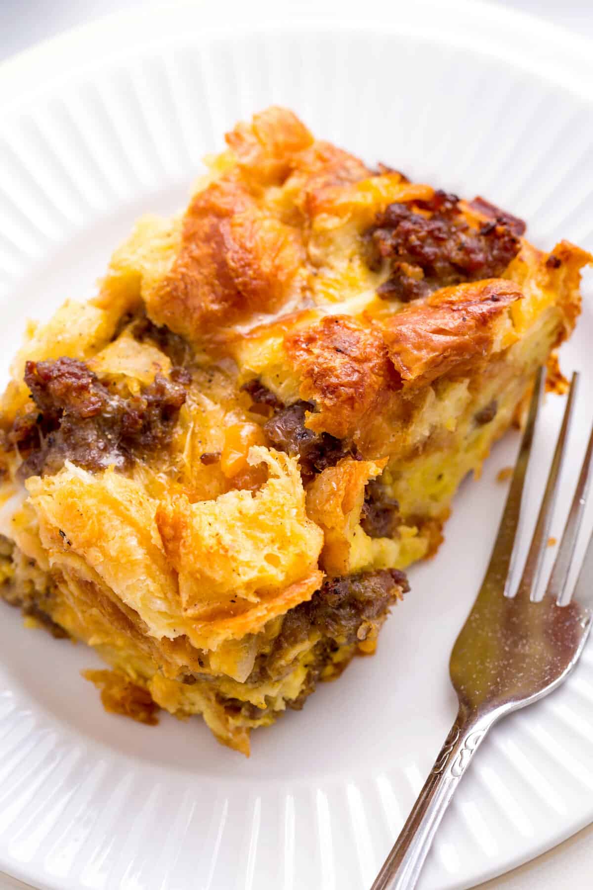 close up image of a serving of sausage croissant breakfast casserole served on a white round plate with a silver fork