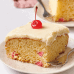 A slice of frosted cherry cake topped with a cherry on a plate.