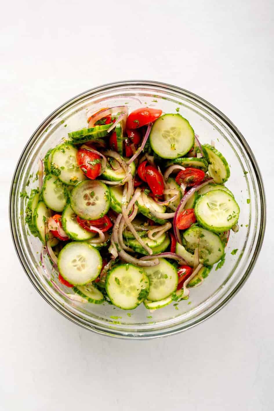 sliced persian cucumbers, cherry tomatoes and red onion in a large glass bowl mixed with dressing