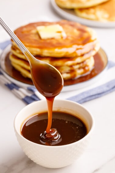 A bowl of pancake syrup with a stack of pancakes in the back.