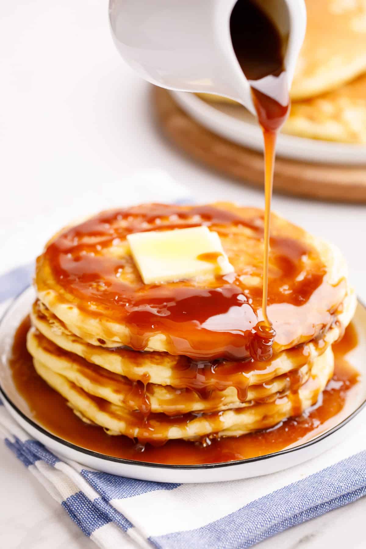 homemade syrup poured out of a vessel over a stack of four pancakes topped with a slab of butter