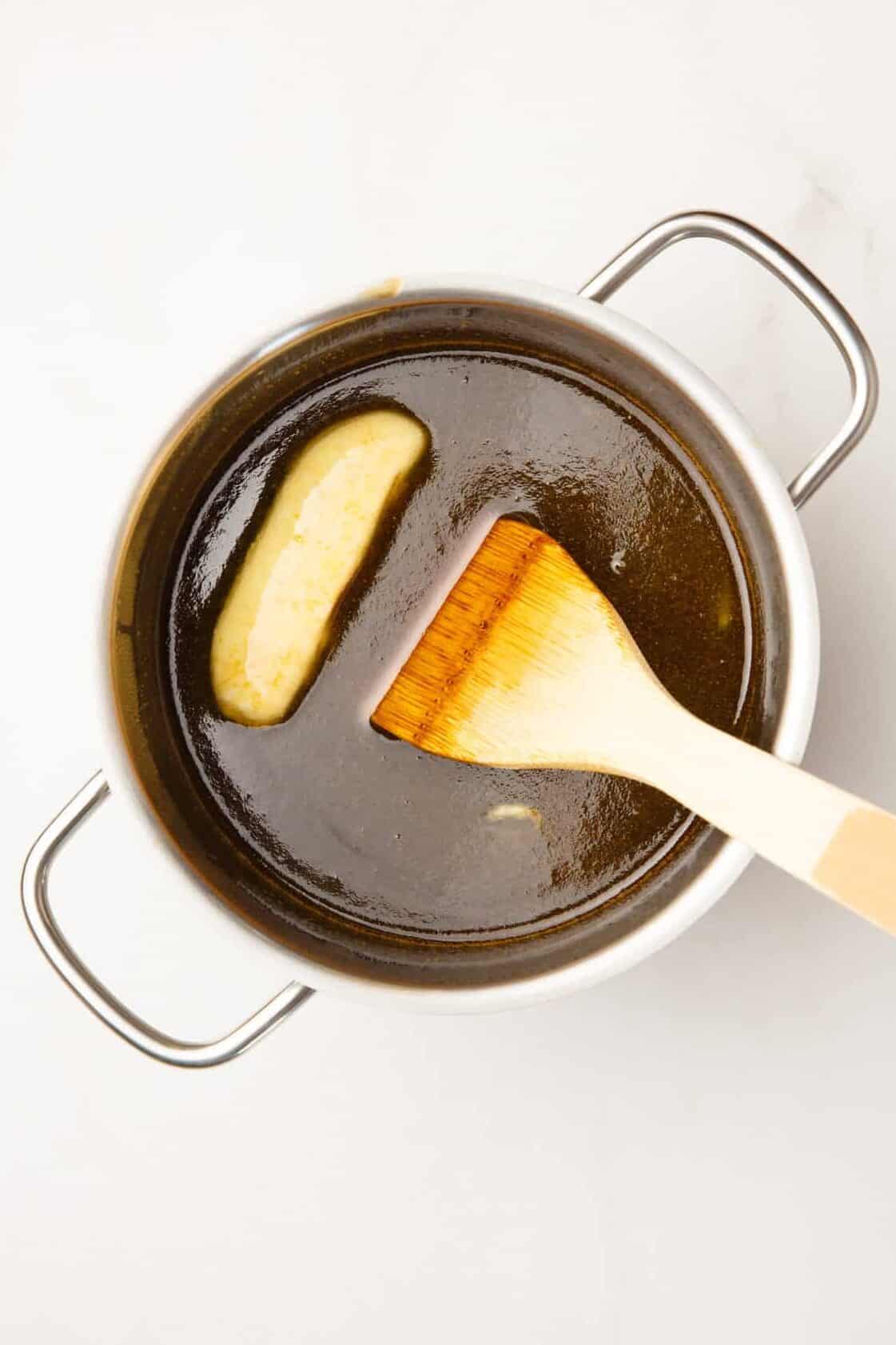 pancake syrup in a stainless steel sauce pan with a stick of butter melting and a wood spatula stirring the pot