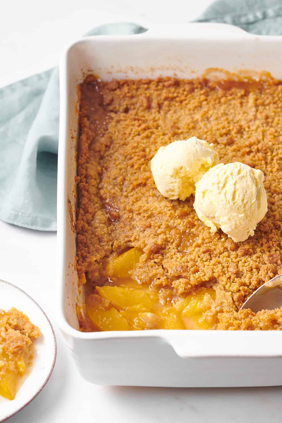 close up image of a casserole dish of peach crumble topped with two scoops of vanilla ice cream
