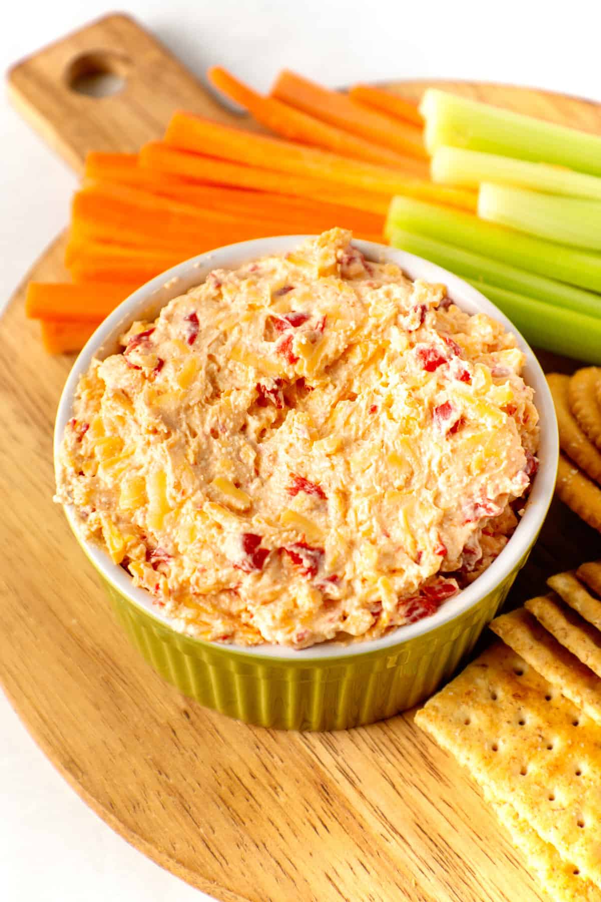 green ceramic bowl with pimento cheese surrounded by a variety of crackers and celery and carrot sticks served on a round wooden board